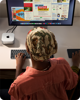 Inimene kirjutab laua taga, ees Mac mini, mis on ühendatud Studio Displayga, Magic Keyboard ja Magic Trackpad ning mitmesugused esemed