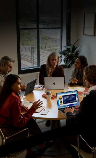 Five individuals seated around a conference table, collaborating on MacBook laptops, with iPhone devices face down on the table