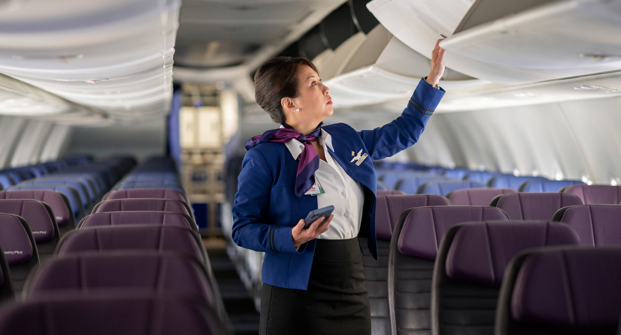 A flight attendant holds an iPhone while opening an overhead bin in an empty aircraft cabin