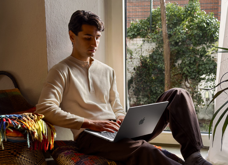 A person sitting on the floor using MacBook next to a window.