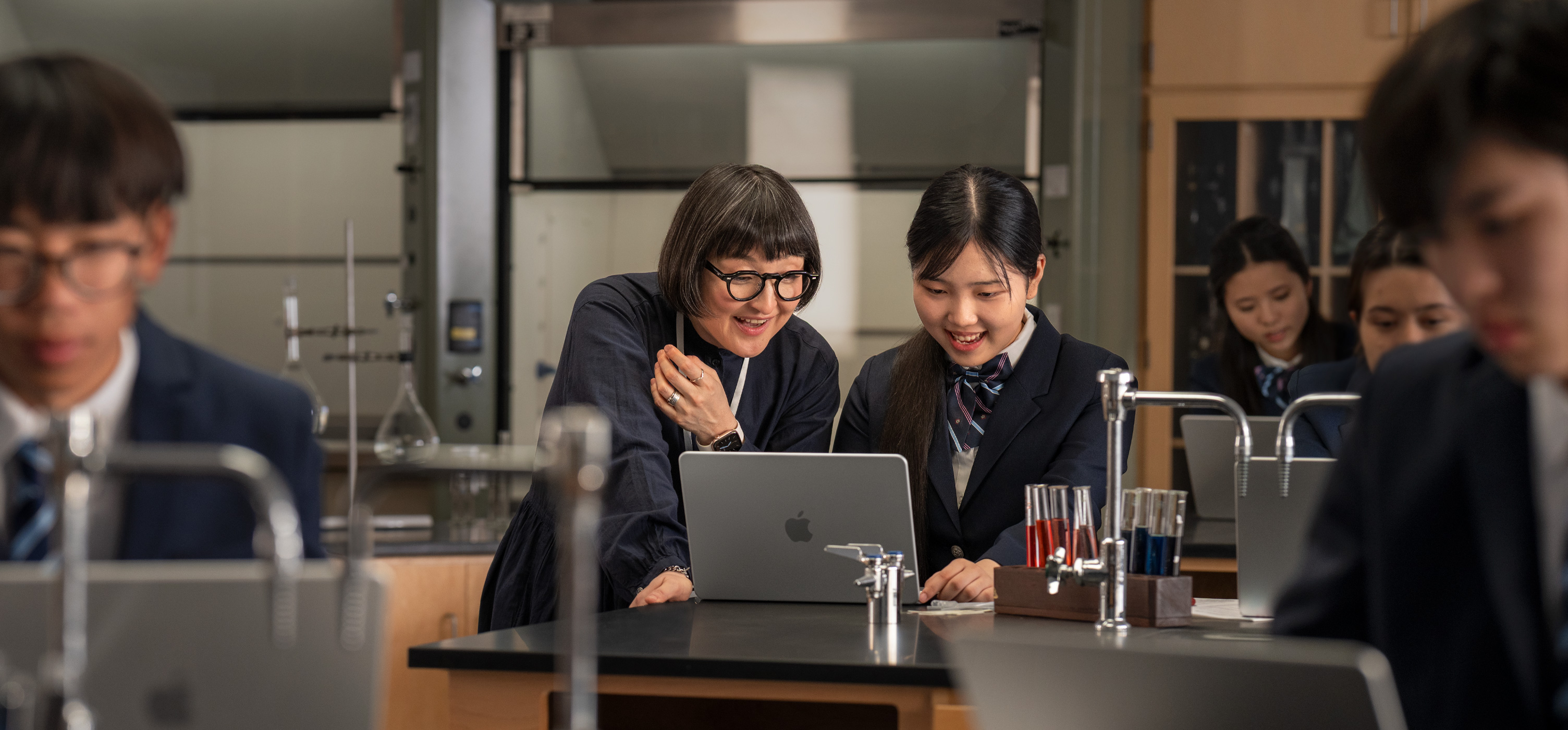 MacBook, open on a table with a student and teacher interacting in laboratory setting, surrounded by test tubes and equipment