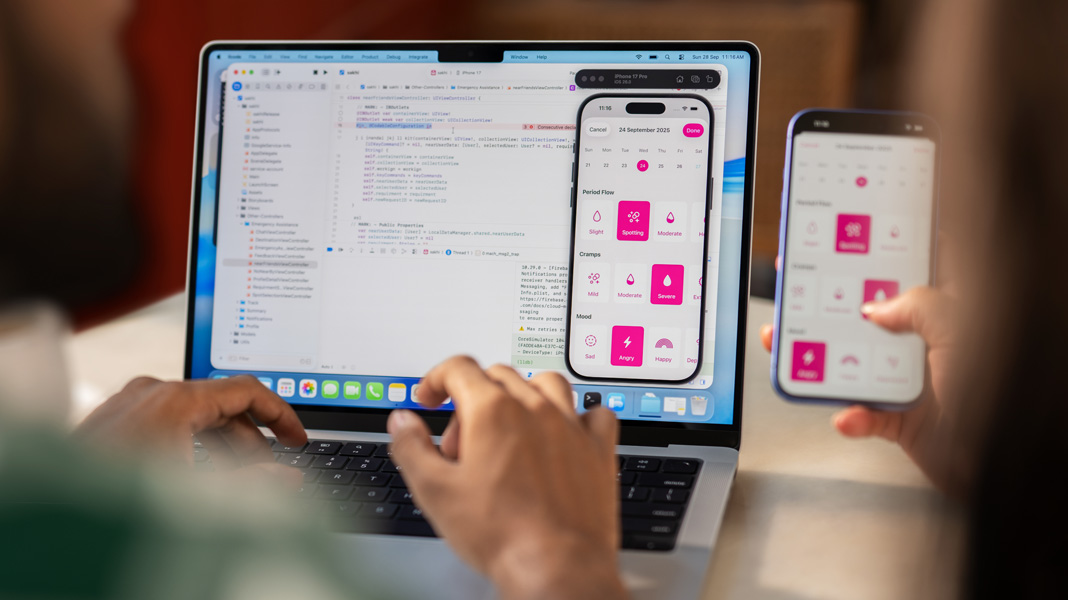 An over-the-shoulder view of a person typing on a MacBook running Xcode, while someone holds an iPhone displaying the Sakhi app, also being shown on the MacBook.