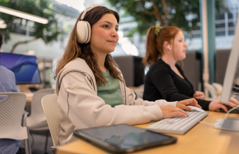 A student wearing AirPods Max sits at a computer typing on a keyboard, with a tablet resting nearby, while another student works next to her.