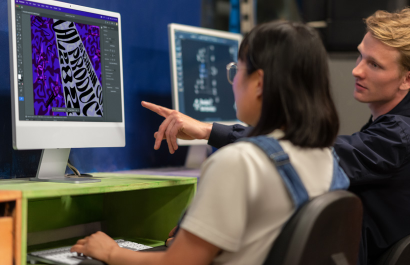 Two students sit in front of iMac monitors displaying colourful models, one pointing at the screen.