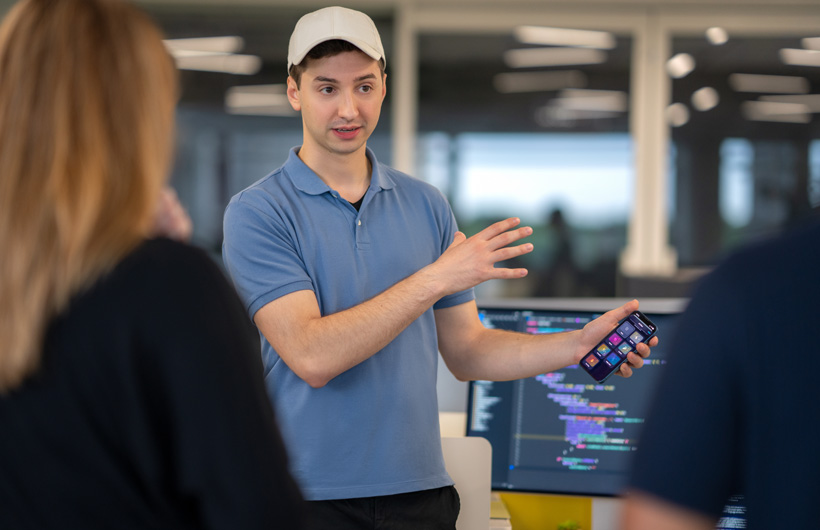 A student speaks to two people and gestures at an iPhone displaying a home screen of apps. Behind him, a monitor shows a code editor with colourful syntax-highlighted text.