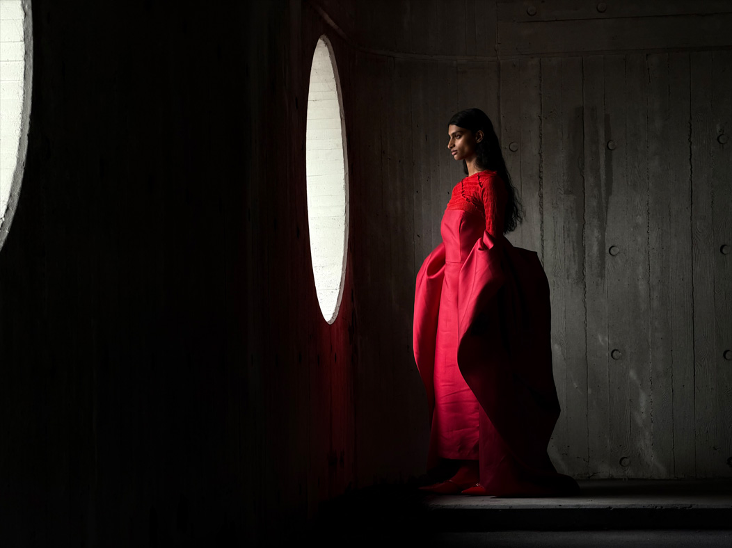 Uma mulher de vestido vermelho longo está sendo fotografada em um ambiente vazio. O app Fotos alterna entre as distâncias focais para demonstrar os recursos do sistema de câmera Pro, mostrando: 100 mm