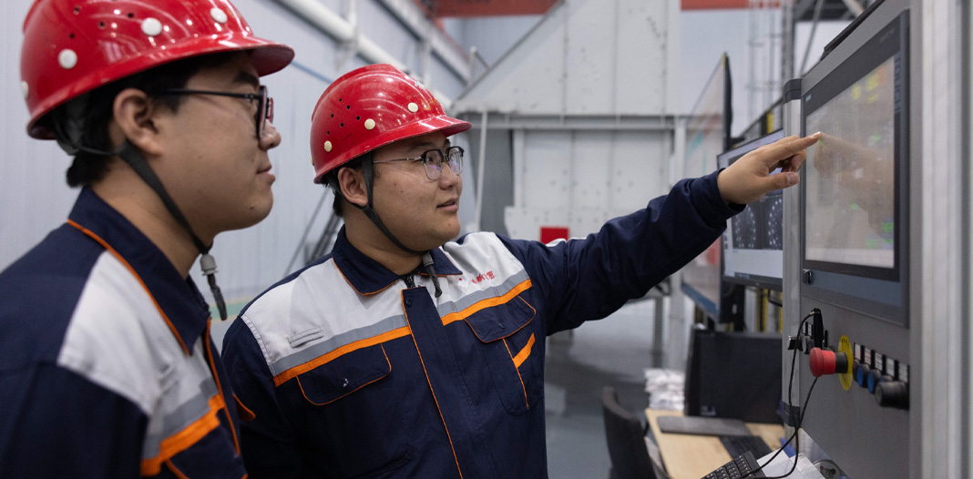 안전모와 안전 유니폼을 착용한 직원 두 명이 하단에 제어 핸들이 있는 디지털 화면을 보고 있는 모습