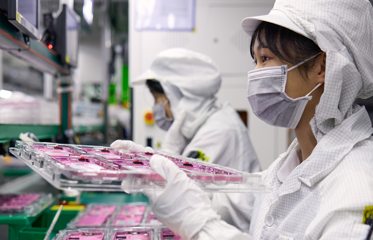 전신 작업복을 입은 직원 두 명이 조립 시설에서 iPhone 부품을 다루고 있는 모습