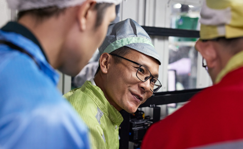 작업 유니폼을 입은 직원 세 명이 AirPods Pro 조립 기계 근처에서 이야기를 나누고 있는 모습