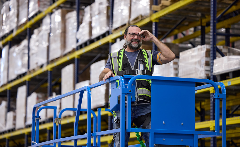 A warehouse employee wearing a safety vest rides a drivable lift